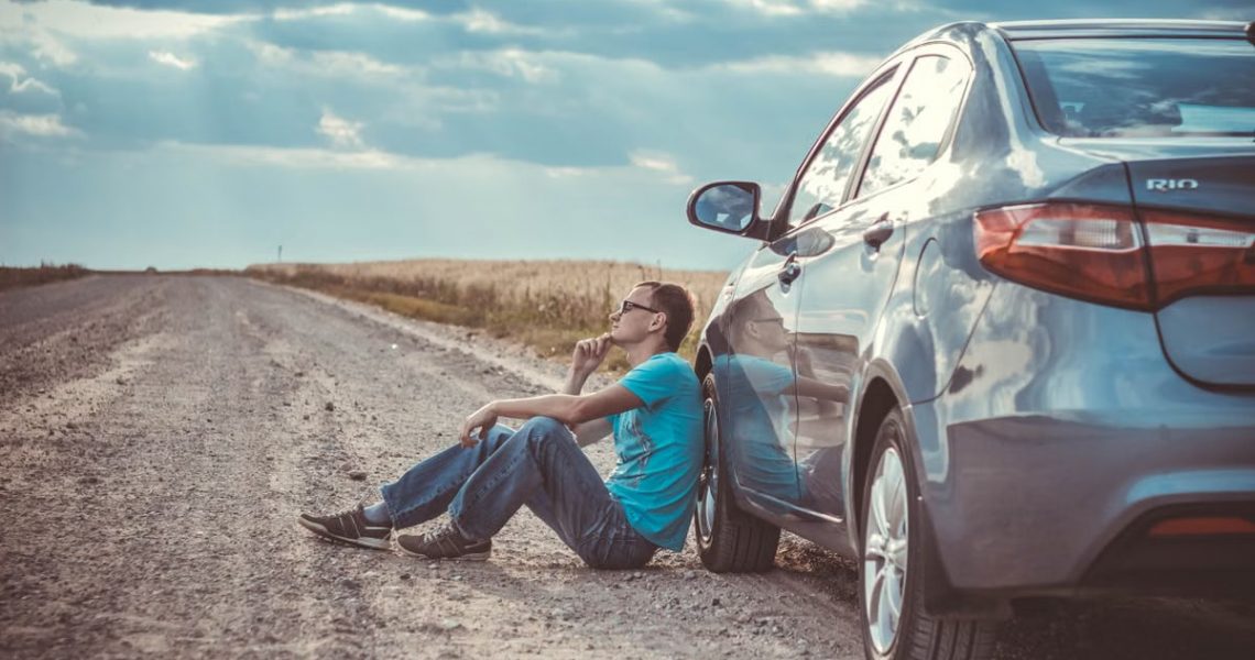 Man sitting by a car on a remote gravel road under a cloudy sky, deep in thought.